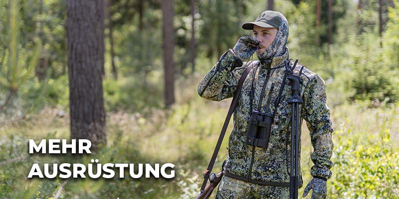 Jäger in tarnfarbener Kleidung steht im Wald und bläst in einen Lockruf; um den Hals trägt er ein Fernglas und auf dem Rücken ein Stativ.